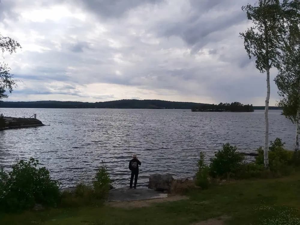 En naturskön vy med ett lugnt vattendrag omgiven av grönska och skogskantade stränder under en molnig himmel, typisk för landskapet vid Hätte Camping.