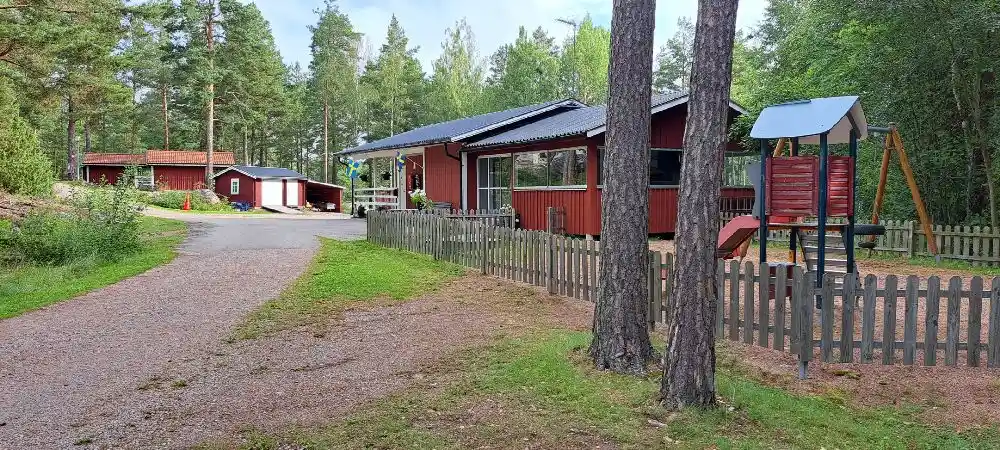 En grönskande campingplats med träd, gräs och buskage omger en liten stuga av trä i en lantlig miljö.