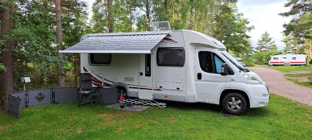 En campingplats med flera husbilar och husvagnar, där en av husvagnarna har en utfälld markis. Vissa fordon har öppna bildörrar och det finns även en lastbil i närheten.