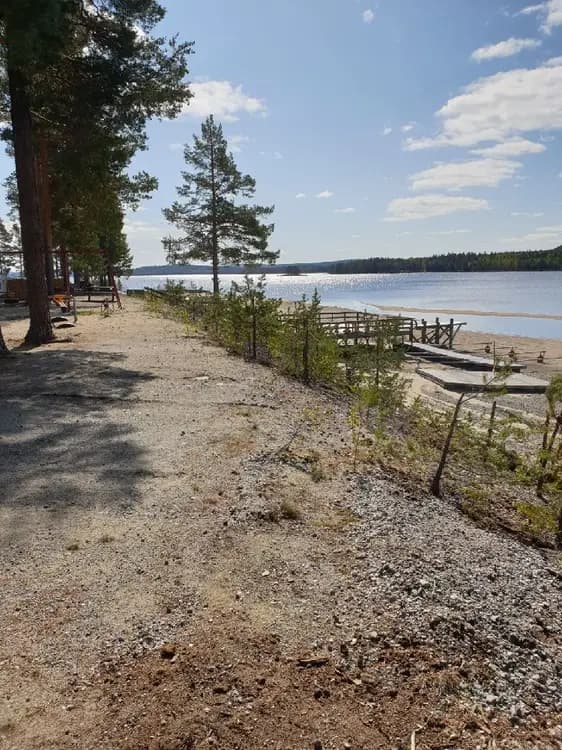 Vy över en naturskön kustlinje vid Sandnäsets bad & camping, med en fridfull strand som möter det glittrande vattnet från en sjö, omgiven av grönskande landskap.