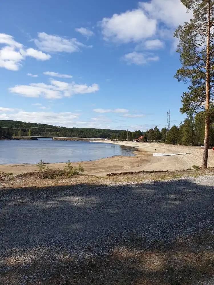 Vy över en sjö med klart vatten och molnig himmel vid Sandnäsets bad & camping, omgiven av en naturskön kustlinje och grönskande stränder.