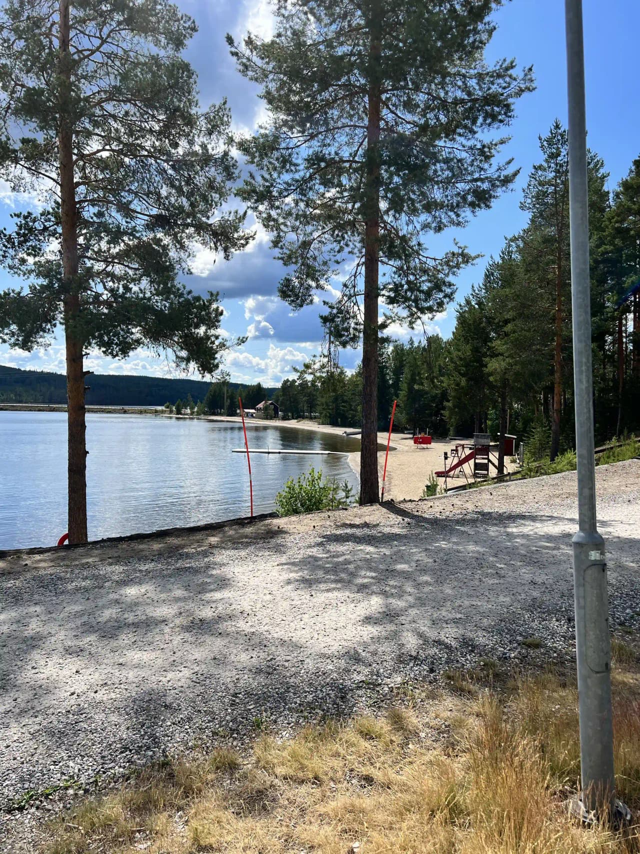 En pittoresk sandstrand vid Sandnäsets bad & camping med utsikt över en stilla sjö, omgiven av grönskande natur och mjuka kullar i bakgrunden.