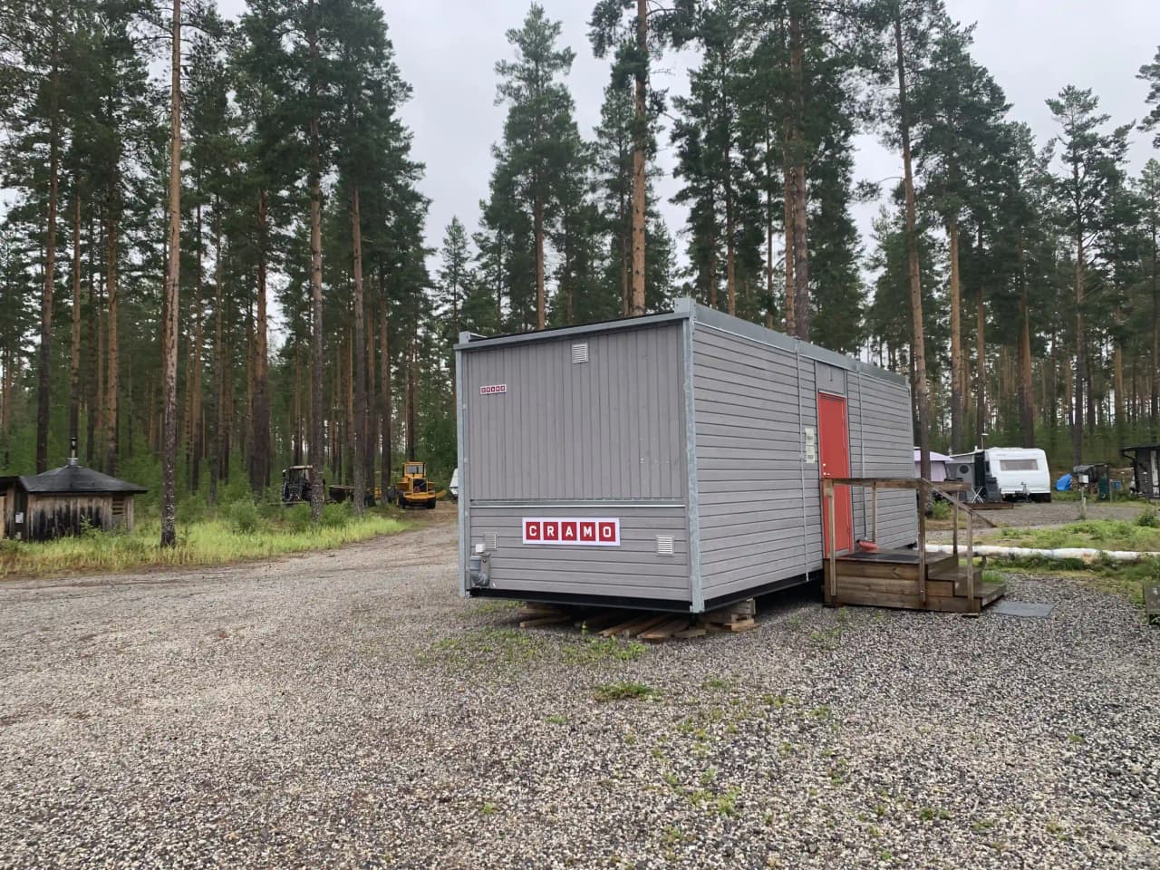 En campingplats vid Sandnäset med flera husvagnar och ett par små byggnader omgivna av lummiga lärkträd. En lastbil och en container syns i bakgrunden.