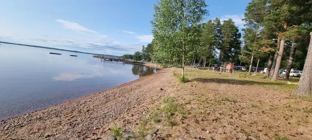En naturskön bild av sjön vid Tällbergs camping, med lugnt vatten och en strandkant, omgiven av grönskande träd och himmel i bakgrunden.