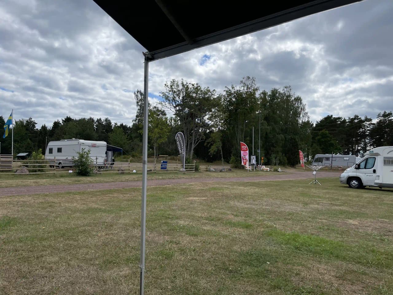En campingplats med husvagnar och husbilar parkerade under skuggande träd. Några tält syns i bakgrunden och en stolpe står nära platsen, vilket skapar en avkopplande miljö för besökare.