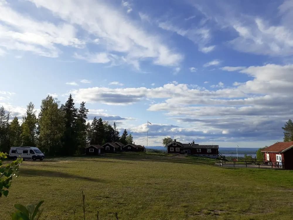 En naturskön bild av Åsengården med gröna ängar och molntäckt himmel, omgiven av ett lantligt landskap med en liten stuga i bakgrunden.