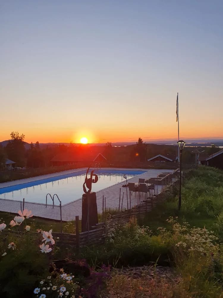 En vy över Åsengården i skymningen, där horisonten lyses upp av solnedgångens varma färger. Besökare njuter av fritidsaktiviteter i den avslappnande och natursköna miljön.