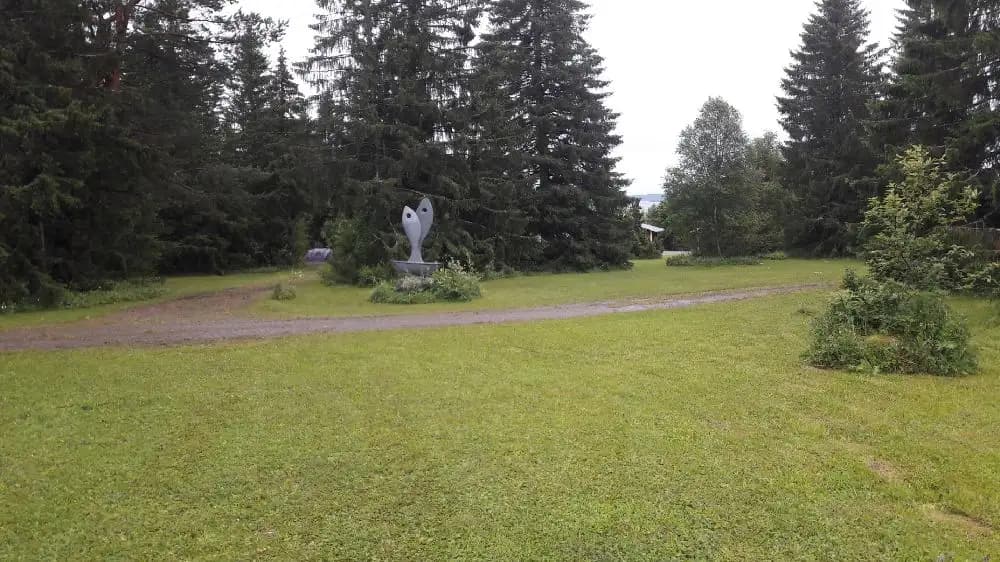 En grönskande campingplats i Ångsta med gräsmattor, buskar och träd i bakgrunden, omgiven av frodig natur och parkeringsarrangemang.