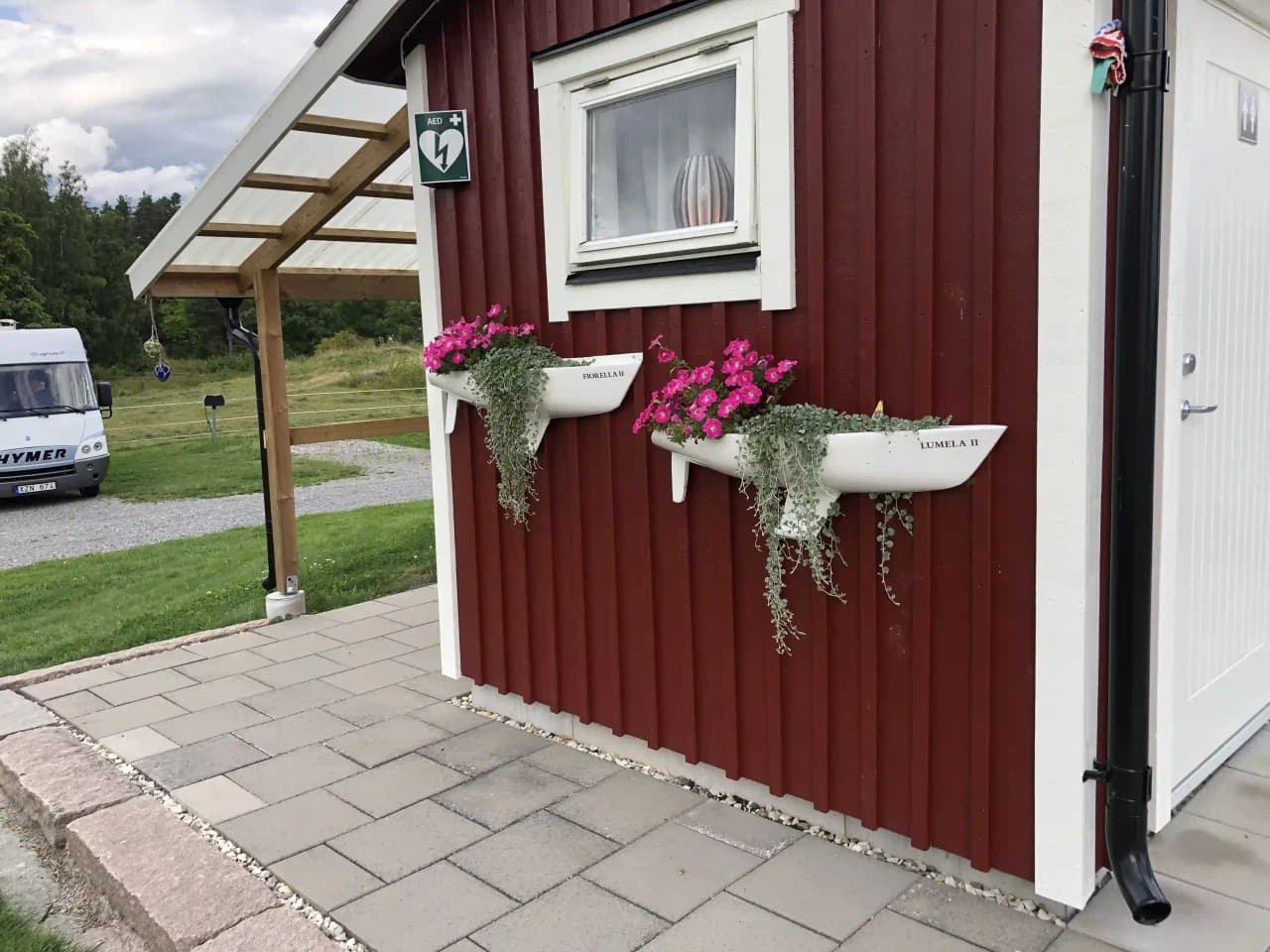 Alt-text: En träbyggnad vid Upperuds Gästhamn, med träpaneler och en trädörr, omgiven av en grönskande bakgård.