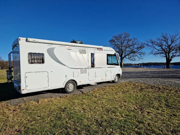 En uppställningsplats vid Upperuds Gästhamn med flera husbilar och husvagnar parkerade. Bakgrunden visar frodig grönska och en delvis klar himmel, vilket ger en trivsam och avkopplande atmosfär i hamnen.