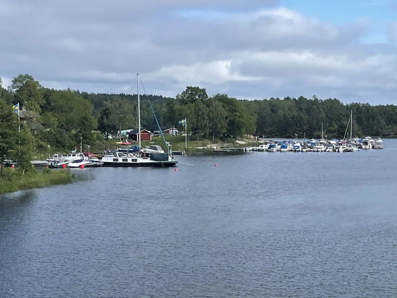 En bild av Upperuds Gästhamn, där flera båtar ligger förtöjda längs kajen i en lugn vattenväg, omgiven av grönska och små kustområden.