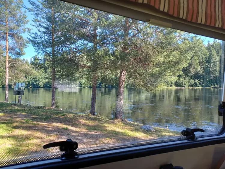 En sjö med spegelblank yta, omgiven av grönska. En smal vattenväg sträcker sig genom landskapet, med träd och buskar som kantar den. En båt syns i bakgrunden, speglad i vattnet.