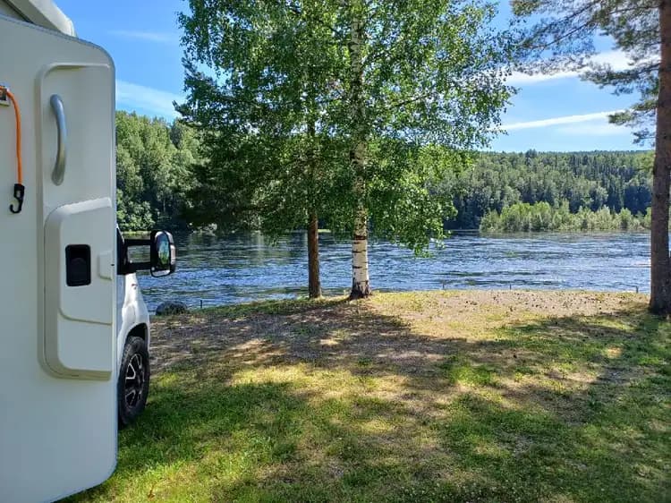 En modern husbil står parkerad vid kanten av en naturskön sjö, omgiven av gröna kullar och skogar. Horrisonterna i fjärran förstärker känslan av frid och äventyr.