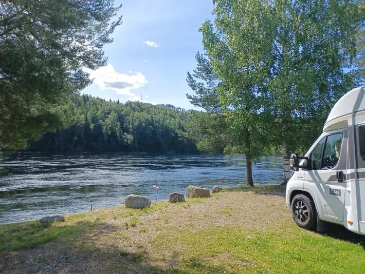 En campingplats vid en sjö omgiven av barrträd, med en husbil parkerad i närheten och ett vattendrag som rinner genom området.
