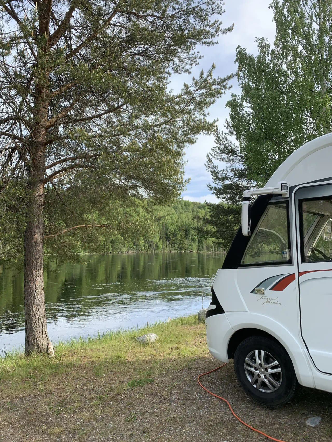 En fritidsfordon står parkerad vid en naturskön sjö med vass och omgivande våtmarker. I bakgrunden syns en båt på vattnet och omgivande grönska, vilket bidrar till en känsla av friluftsliv och äventyr.
