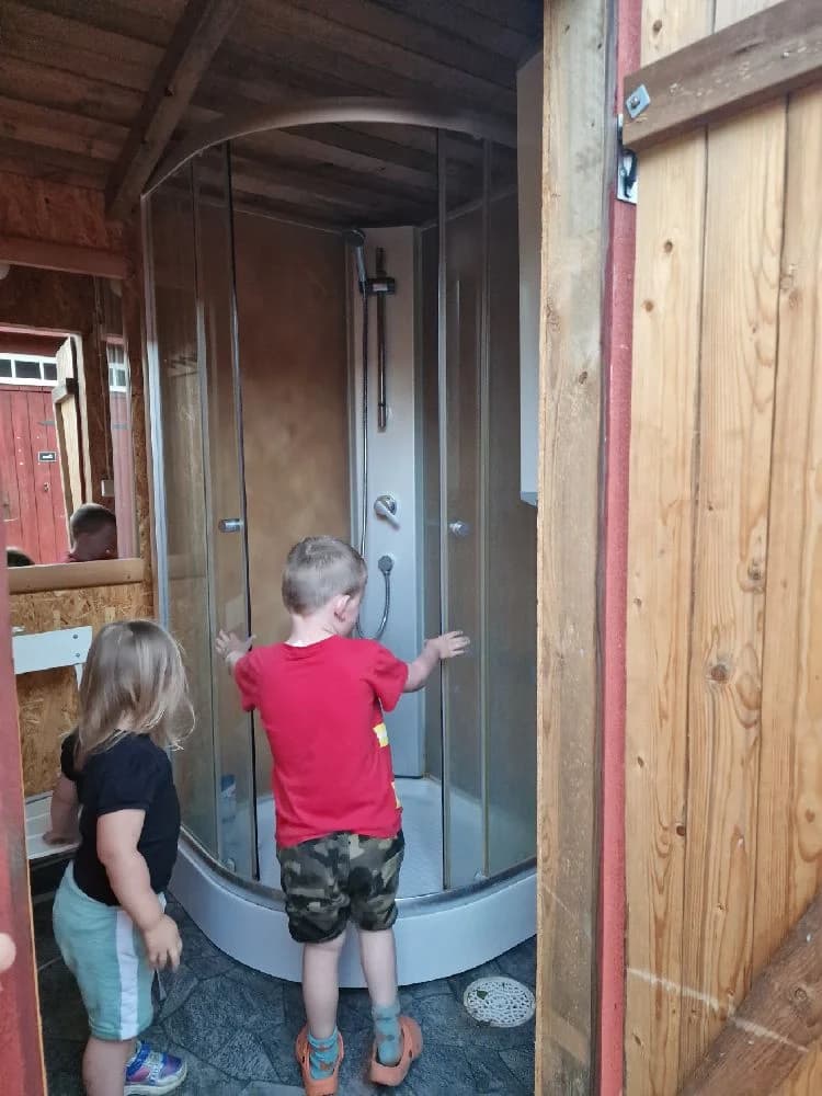 En liten stuga med träpanel och en öppen träport, där ett litet barn står på tröskeln till dörren. Placeringen antyder att det är en del av Mackmyra bruk och golfområde, och omgivningen ger känslan av en rustik och naturnära miljö.