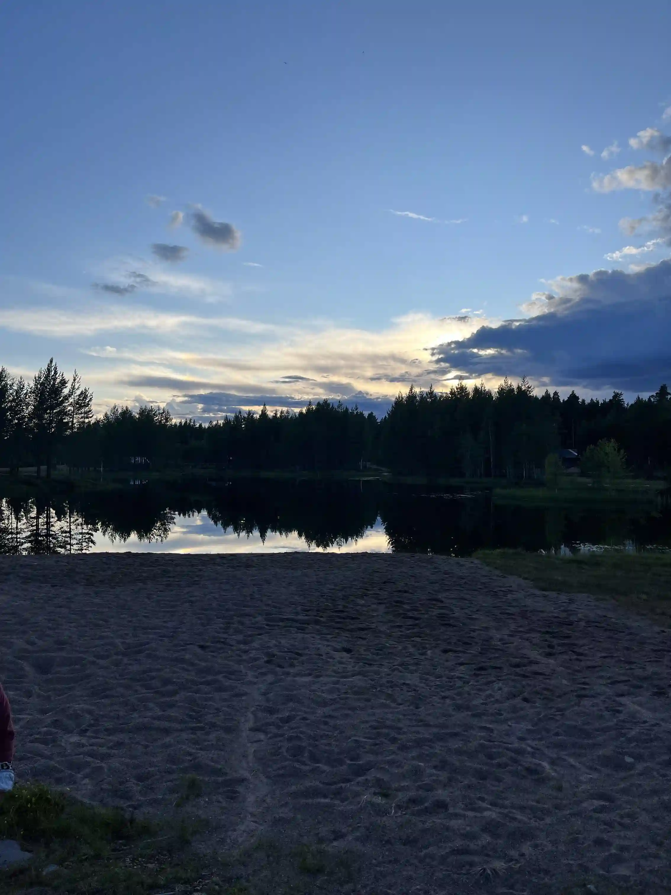 En vacker solnedgång över en sjö i Mora, där himlen är delvis täckt av moln och reflekteras i det stilla vattnet. I bakgrunden syns horisonten klart mot den färgrika kvällshimlen.