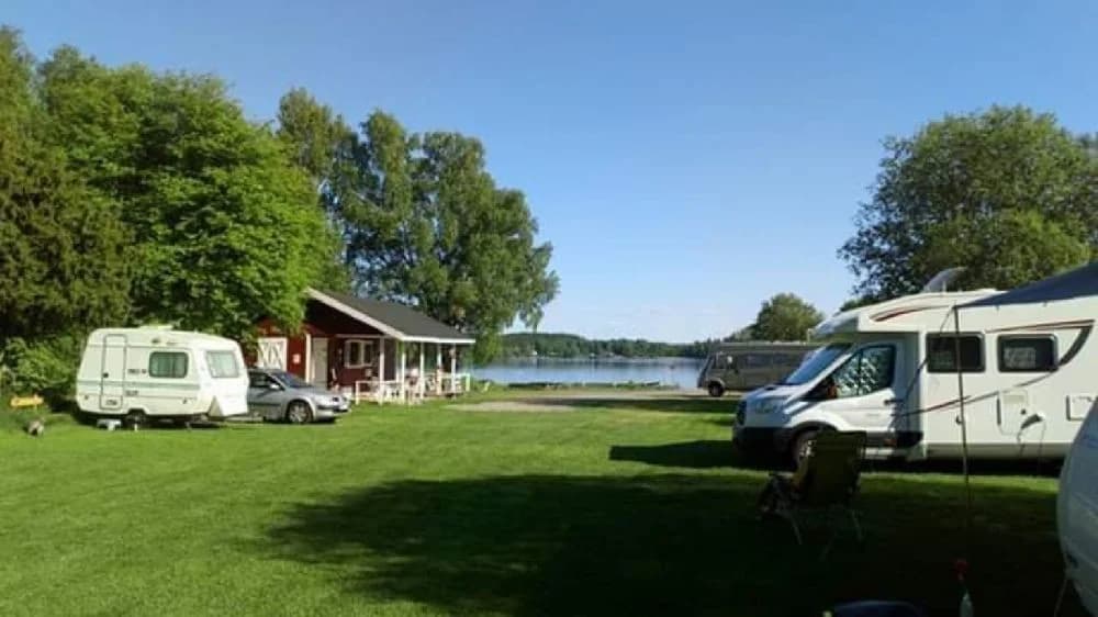 En campingplats med husvagnar och tält uppställda i en grönskande park vid en sjö, med café och rekreationsmöjligheter i närheten.