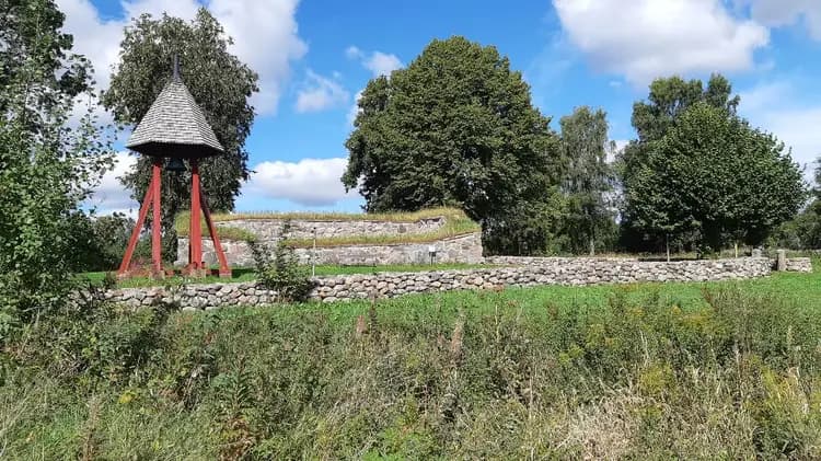 En grön campingplats med välskött gräsmatta och stenmur, omgiven av äng och parkeringsområde.