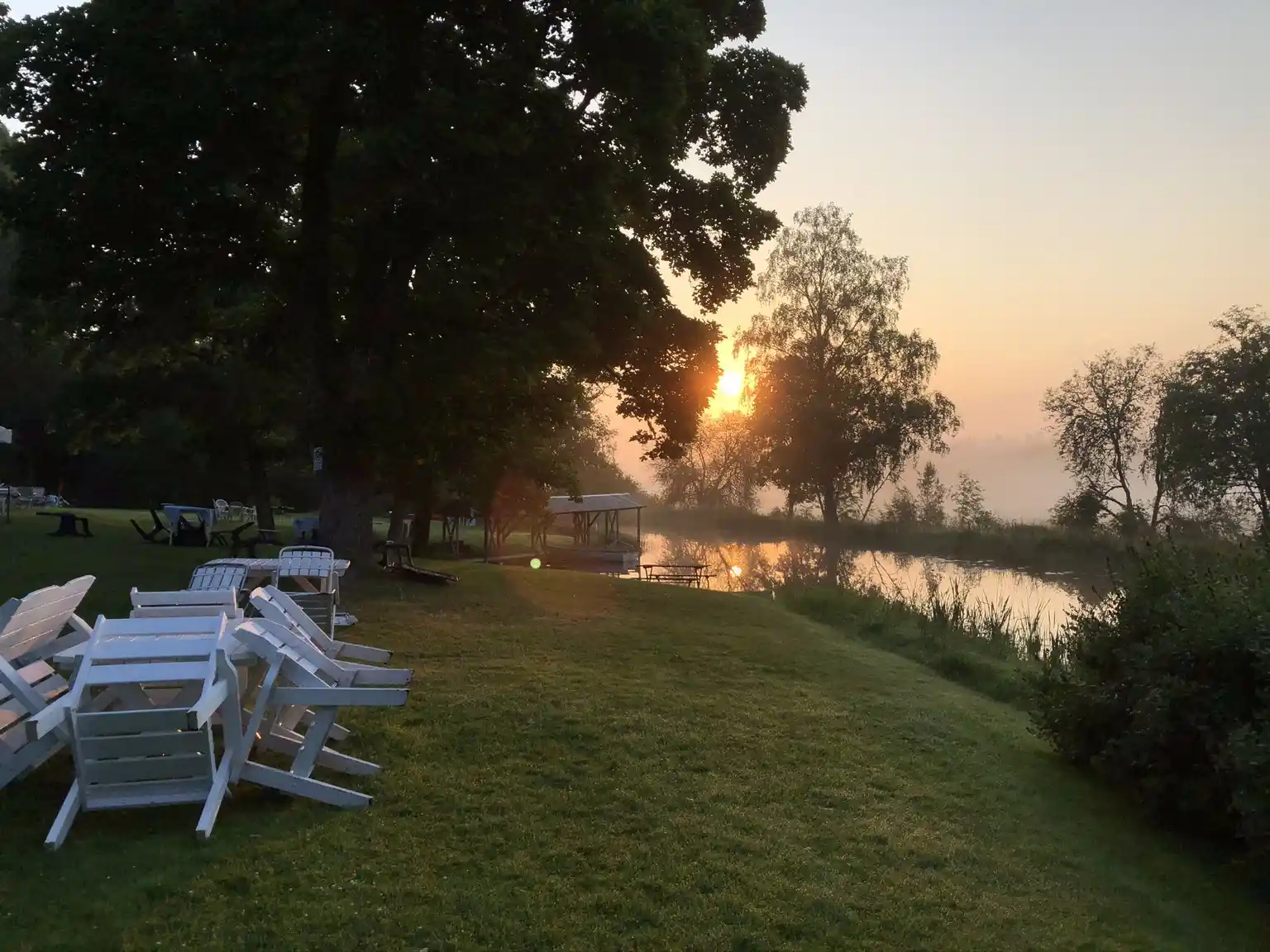 En scen från Badsta Camping med en sjö som speglar de omgivande träden, himlen och landskapet. Det är tidigt på morgonen eller sent på kvällen, och stämningen i bilden är lugn och rofylld. På stranden finns utomhusmöbler som inbjuder till avkoppling i denna natursköna miljö.