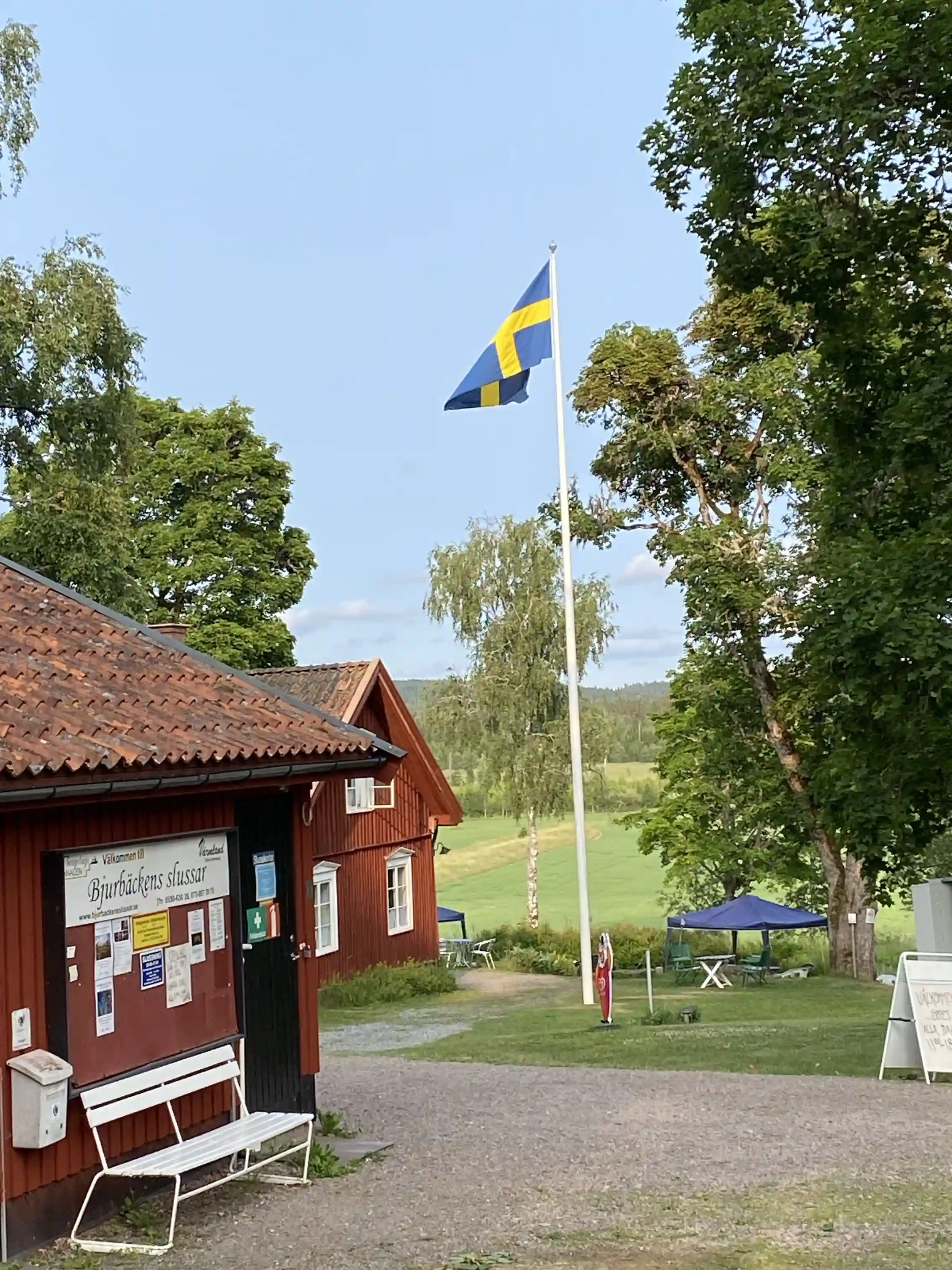 En öppen campingplats med en flaggstång och en flagga vajande i vinden, omgiven av grönska och med fritidsaktiviteter i närheten.