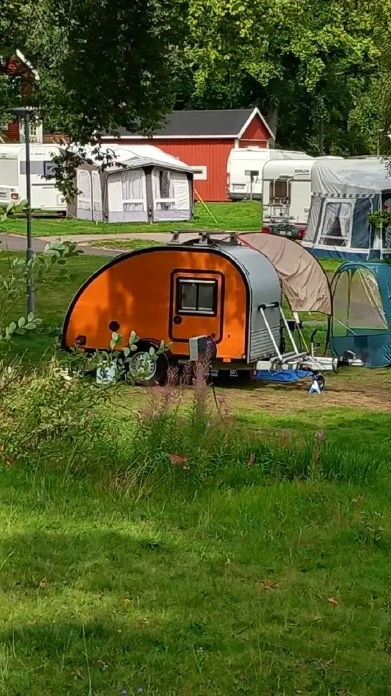 En grön campingplats med tält och husvagnar utspridda över en gräsmatta. Några träd och en lastbil kan ses i bakgrunden, vilket ger en känsla av frihet och friluftsliv på Evedals Camping.