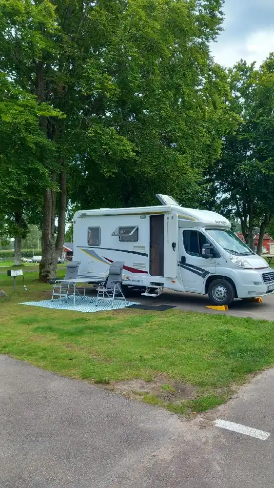 En stor campingplats med flera husvagnar och husbilar parkerade på en välskött gräsmatta. Området är omgivet av träd och ger en lugn atmosfär. Flera fordon och campingutrustning syns också, vilket ger en känsla av aktivitet och friluftsliv.