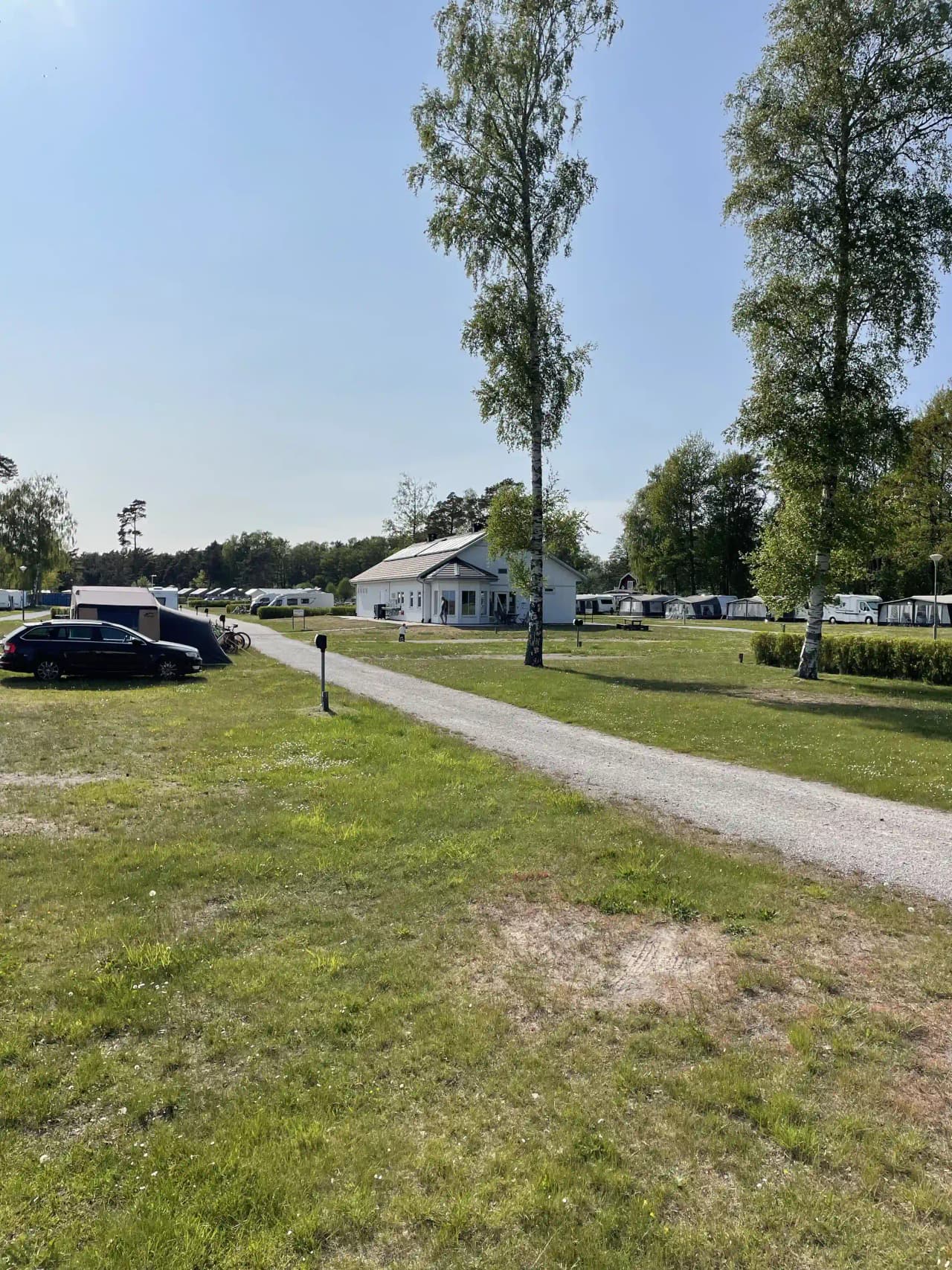 En klar himmel prydd med några moln över en öppen landskap vid Sudersand Resort. I förgrunden syns en grön gräsmatta, parkerade fordon och ett rekreationsfordon, omgivna av en stor parkeringsplats.