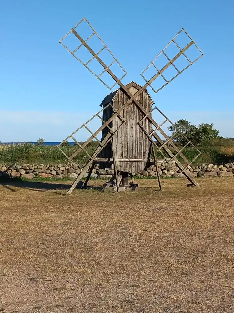 En väderkvarn står på en gräsbevuxen äng under en klarblå himmel vid Wikegårds Semesterby & Ställplatscamping, omgiven av vidsträckta fält och landsbygd.