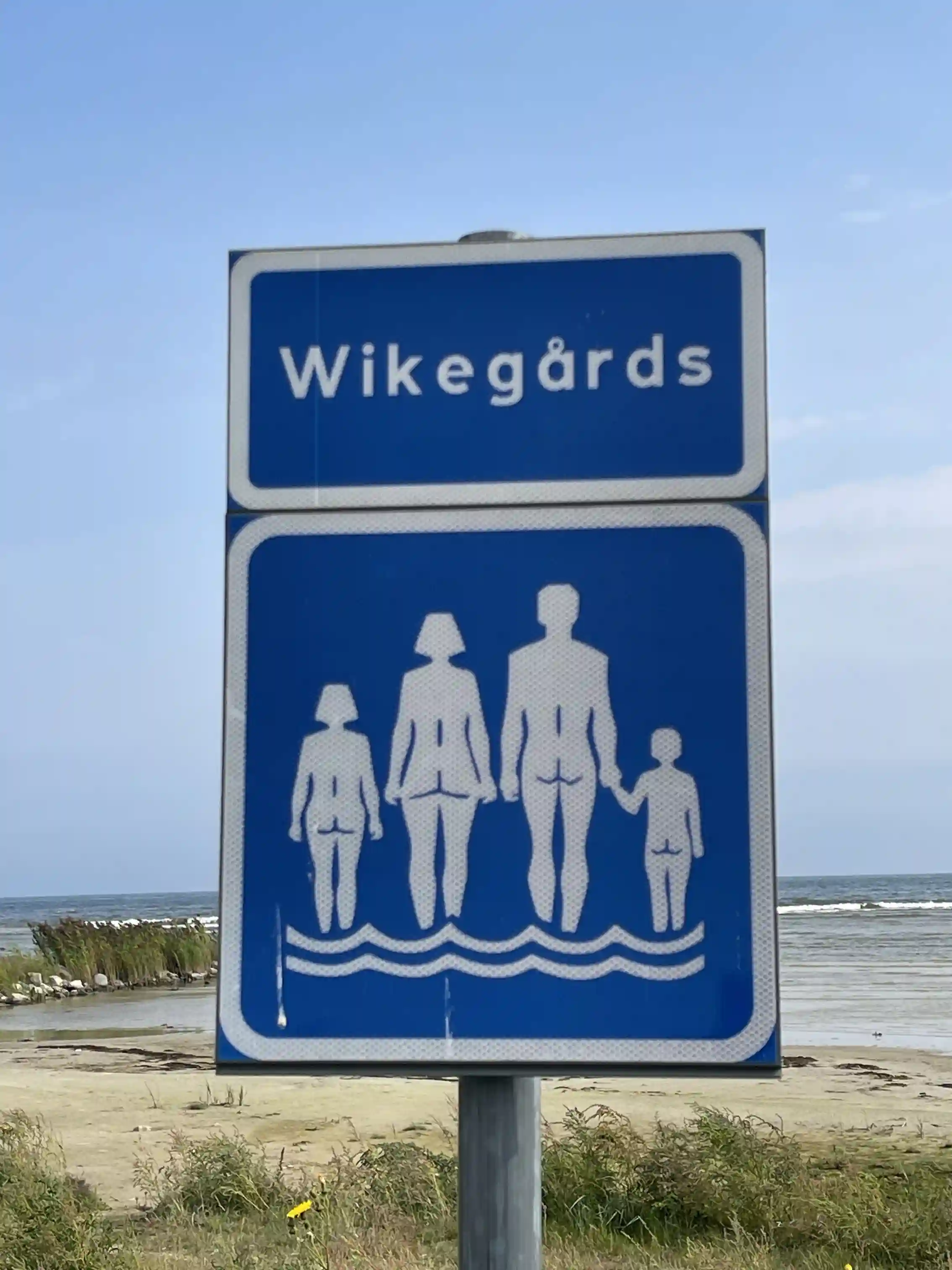 En träskylt markerar ingången till Wikegårds Semesterby & Ställplatscamping omgiven av frodig grönska och promenadvänliga stigar.