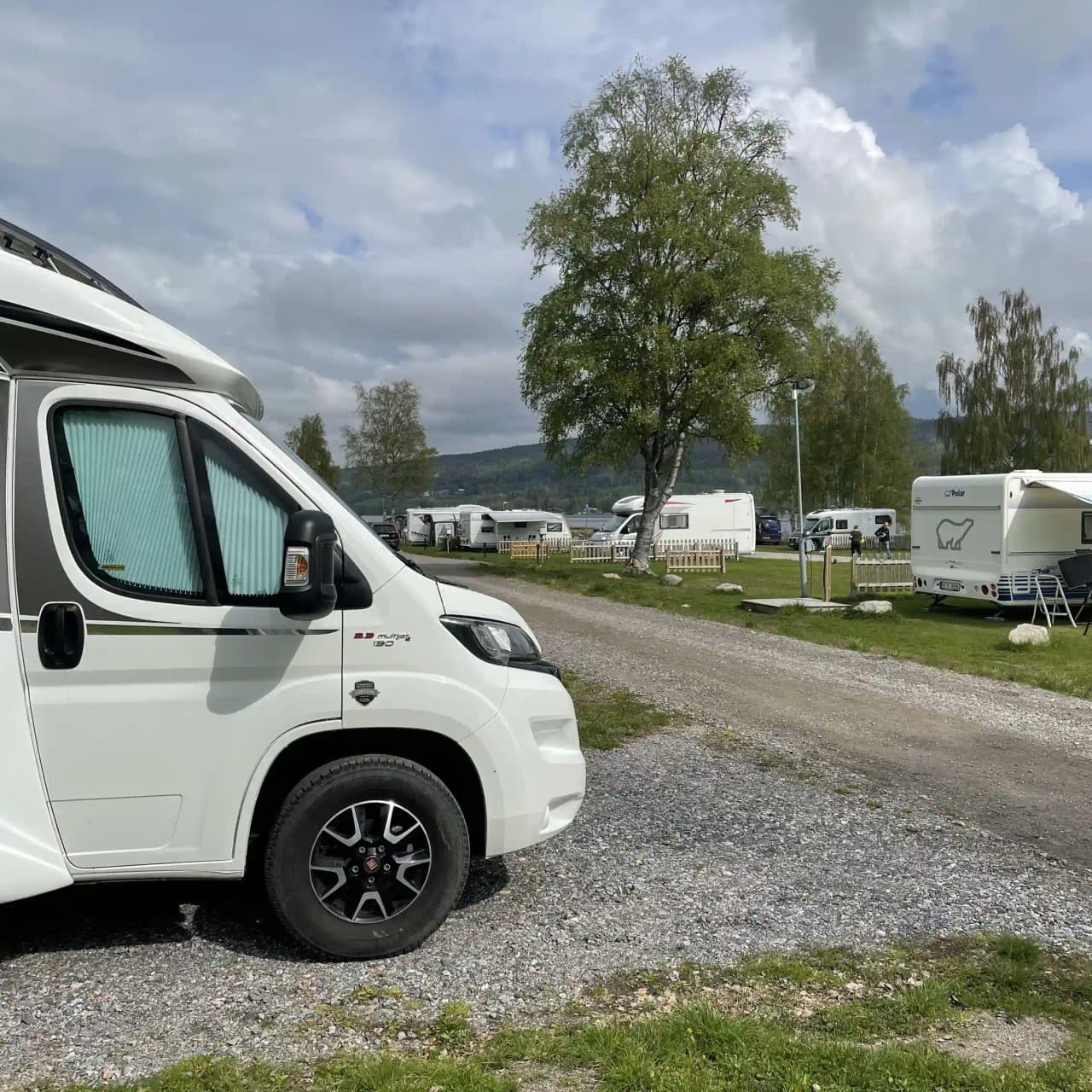 En campingplats med flera husbilar och husvagnar parkerade. Enhetliga fordon står uppställda på grusade områden omgivna av grönska.