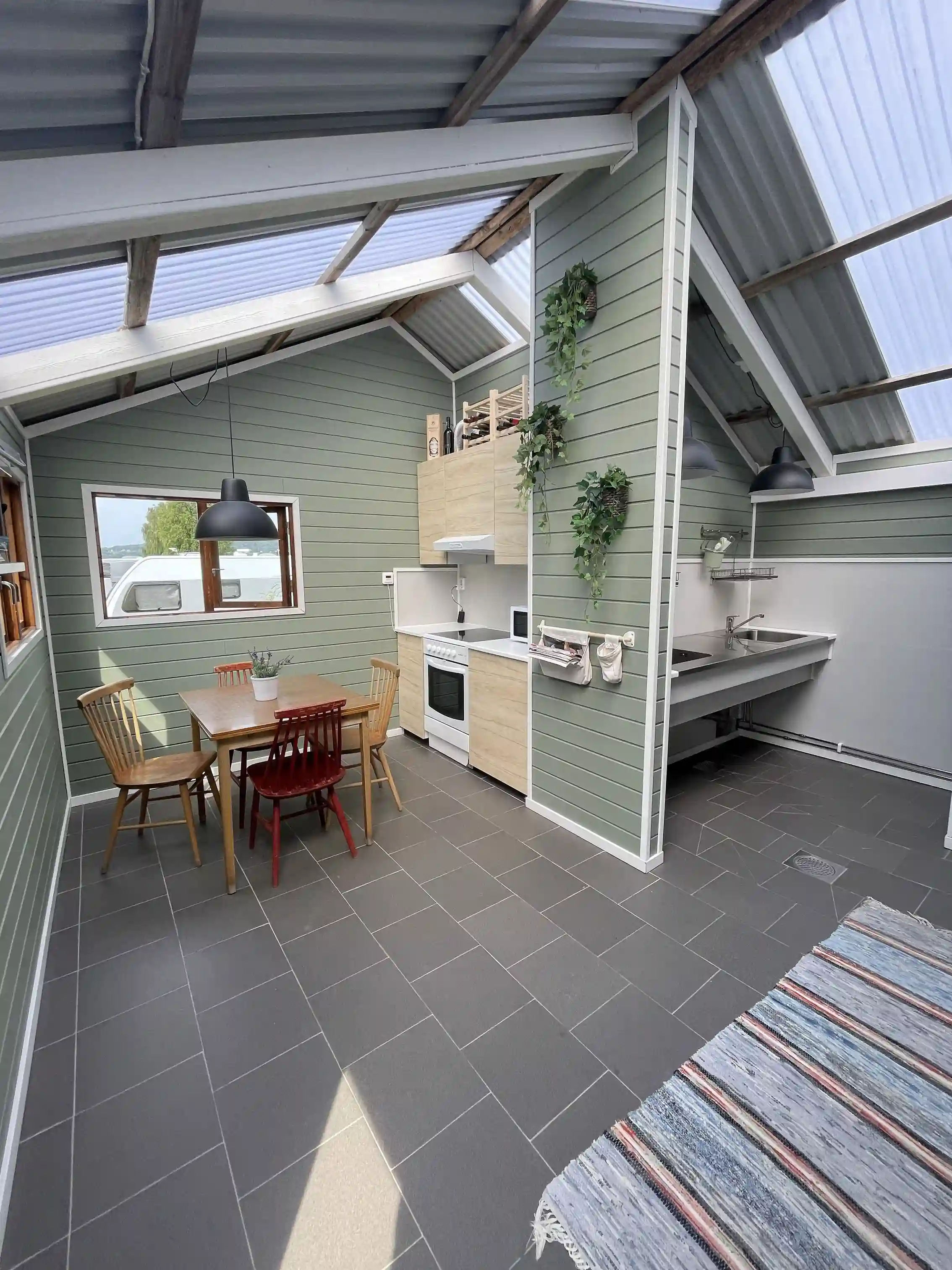 En öppen träpaviljong med tak och väggar av träplankor omgiven av grönska, inredd med stolar och blomkrukor. Ljuset strömmar in och skapar en lugn atmosfär.