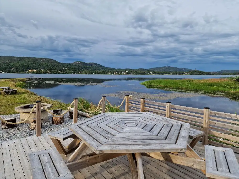 Bild av Overhörnäs camping med en storslagen landskapsscen. Moln svävar ovanför gröna kullar som omger ett glittrande vattendrag. En liten brygga sträcker sig ut i sjön, omgiven av kustnära former.