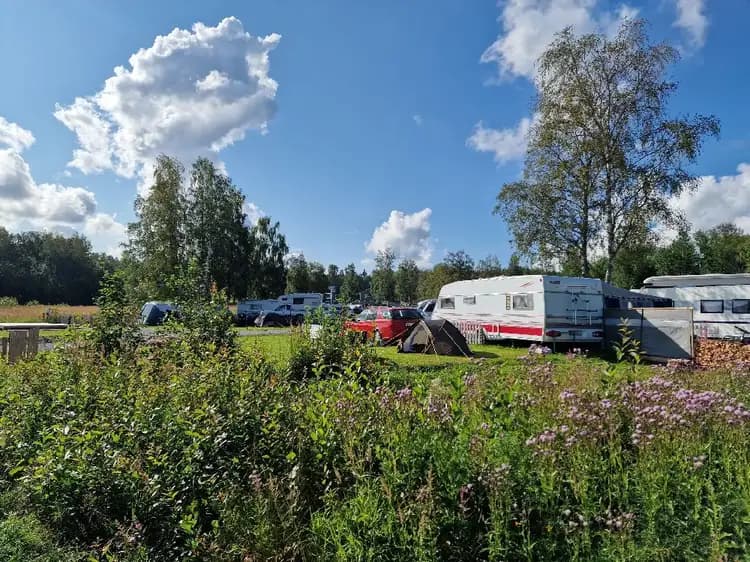 En campingplats med gräs i förgrunden och flera husbilar och husvagnar uppställda. Moln syns på himlen ovan och fordonen står parkerade på en öppen yta.