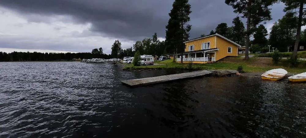 Bilden visar en naturskön vy över en lugn vattenväg omgiven av grönska vid Kolgårdens stugby & camping. I förgrunden syns en liten brygga vid strandkanten.