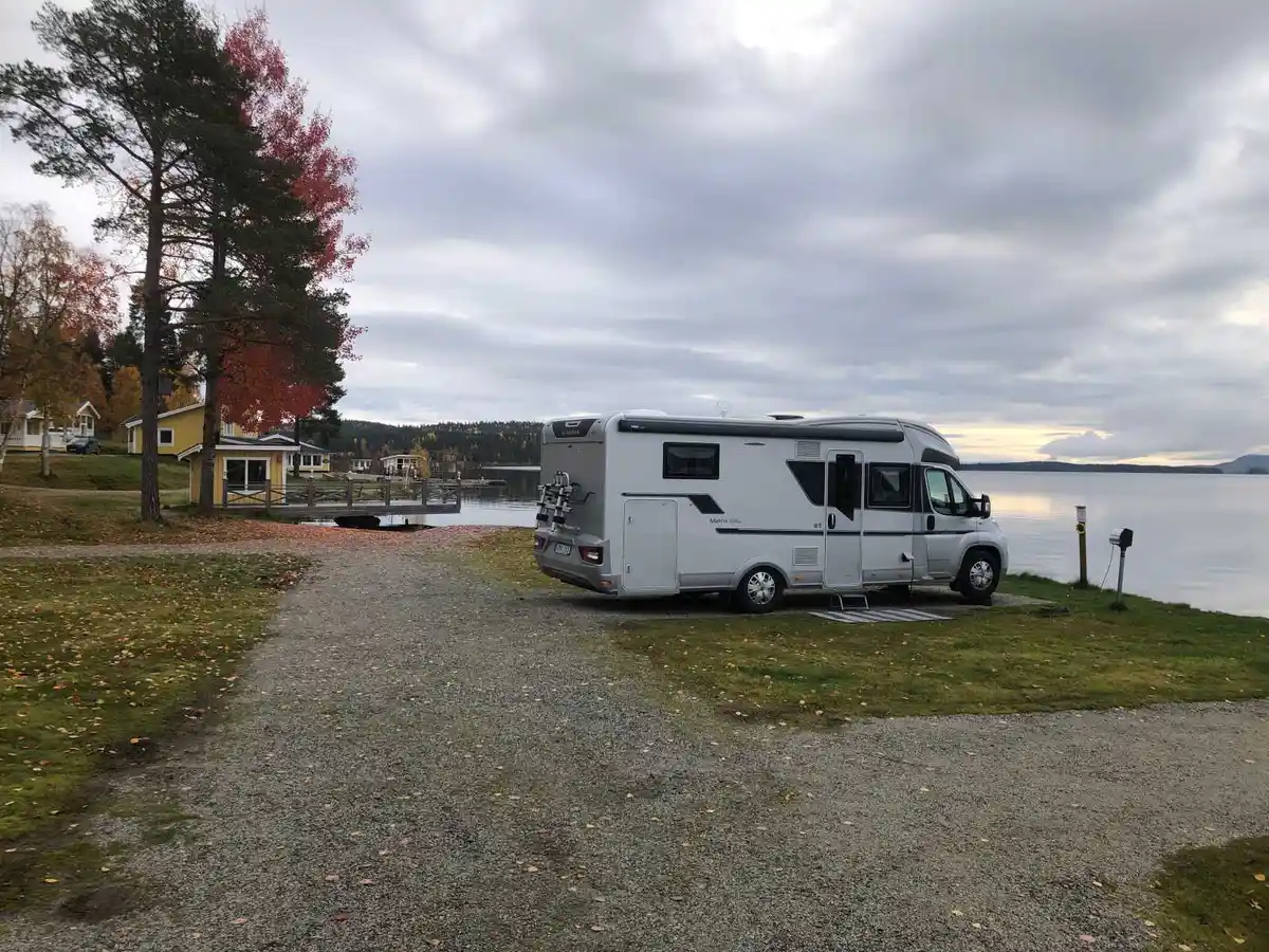 En husvagn står uppställd på Kolgårdens Stugby & Camping, omgiven av lummig natur och nära en sjö. I bakgrunden syns fler campingplatser i det natursköna området.