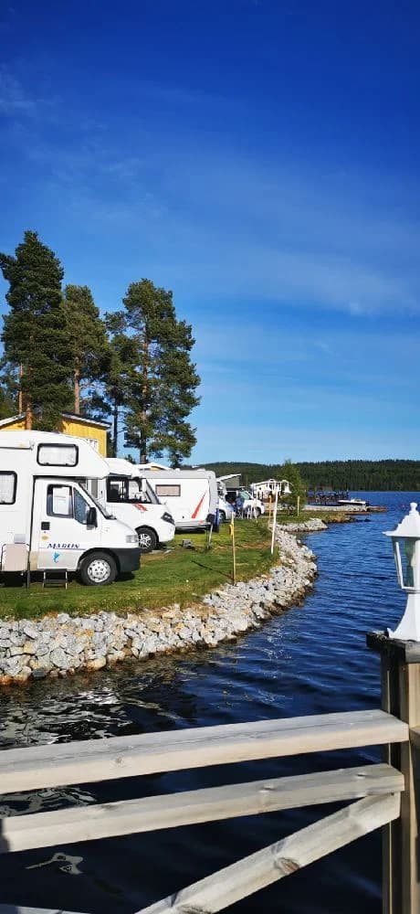 En bild av Kolgårdens Stugby & Camping med en vattenväg och kustlandskap i bakgrunden. I förgrunden syns ett fritidsfordon parkerat vid stranden av en sjö eller hav.