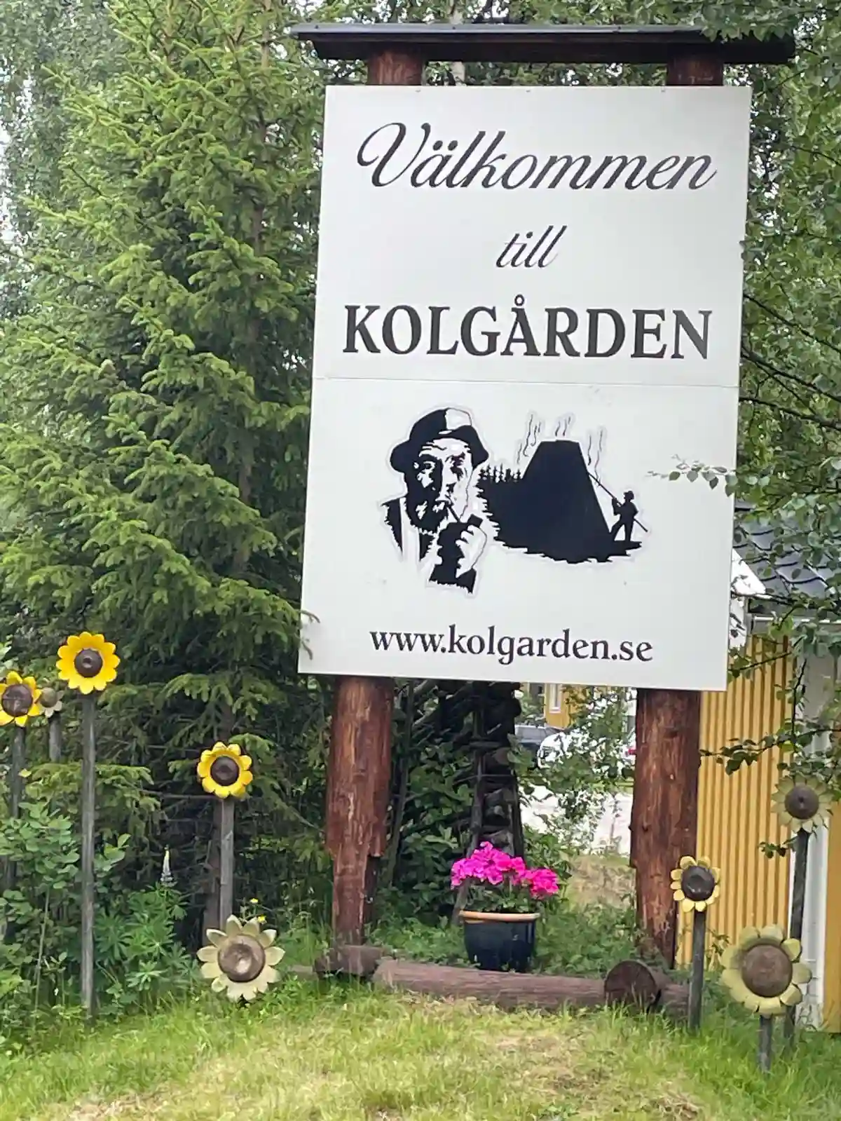 Alt-text: En skylt i Kolgårdens stugby & camping med omgivande blomsterrabatter, solrosor och grönska, som välkomnar besökare.