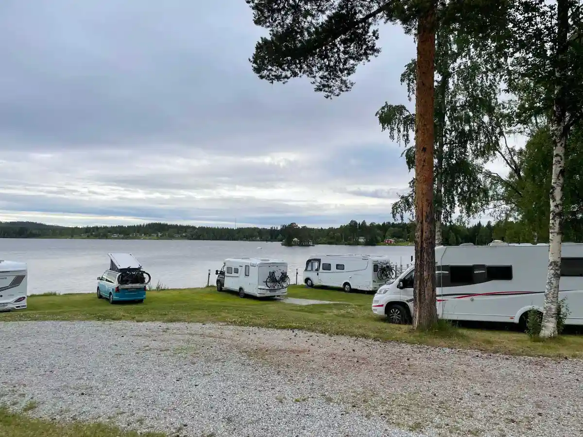 En campingplats vid en sjö med husvagnar och husbilar parkerade. Skogsklädda kullar omger vattnet, vilket ger en naturskön och avkopplande atmosfär.