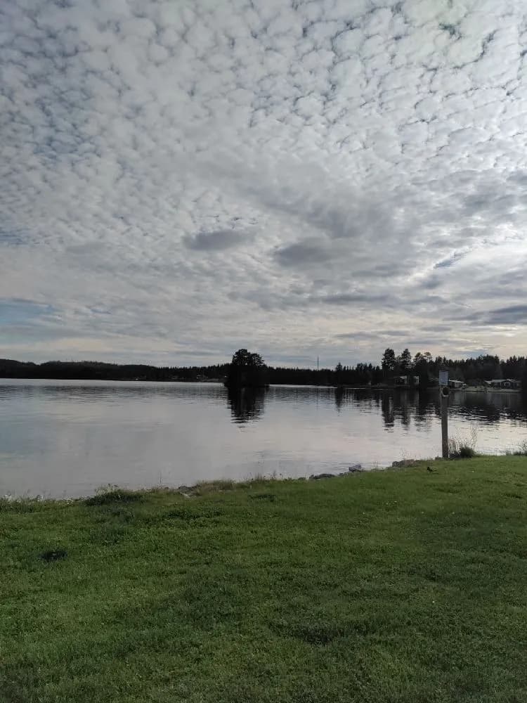 En vy över en sjö med spegelblankt vatten under en klarblå himmel med några molntussar. Naturen runt sjön ger en känsla av lugn och avkoppling.