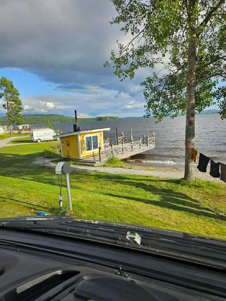 Bild av Kolgårdens stugby & camping: En naturskön vy med en kustlinje som sträcker sig längs en sjö eller ett vattendrag. I förgrunden finns en grön gräsmatta, och i bakgrunden syns molntäckta himmel och omgivande landskap med vatten och skog.