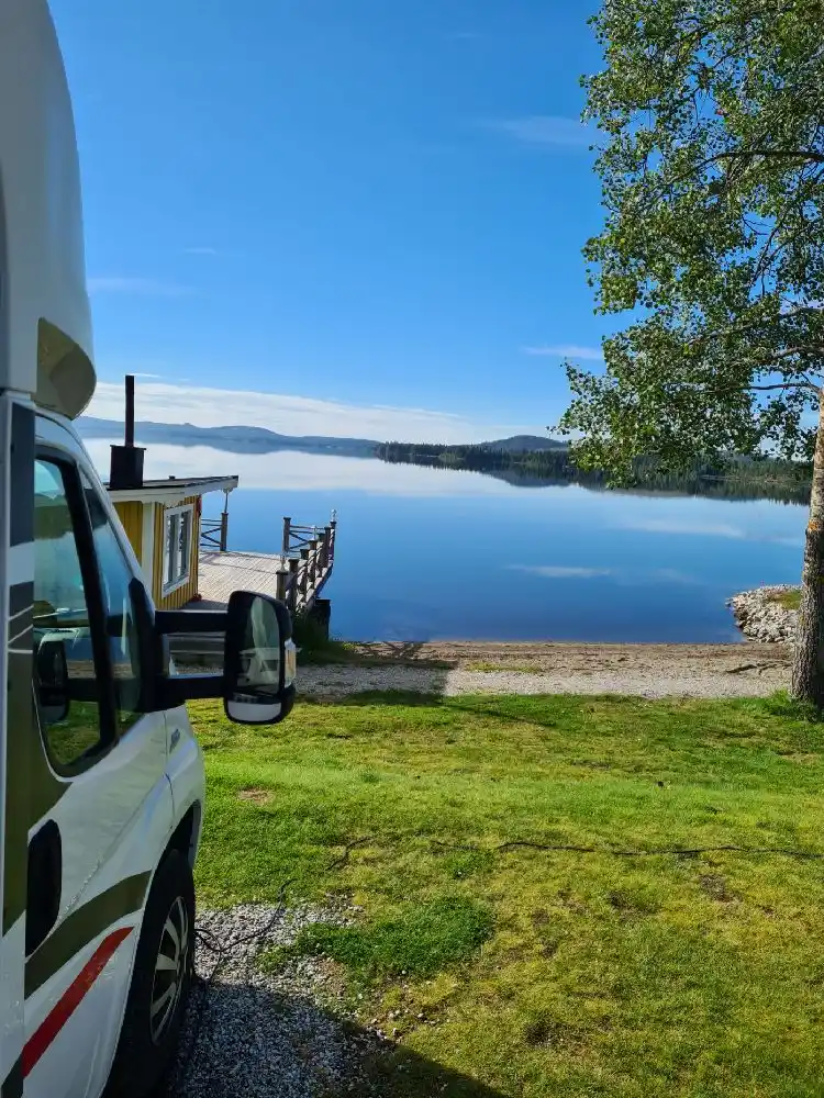 En grön gräsmatta med husvagnar och husbilar parkerade. En picknickbänk står på området nära en vacker sjö med skogbeklädda kullar i bakgrunden vid Kolgårdens stugby & camping.