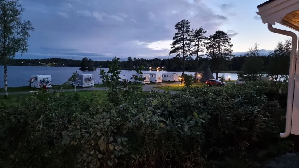 En kvällsbild av en grön gård vid Kolgårdens stugby & camping, omgiven av skuggor och skymningsljus, med en känsla av en sjö i närheten.