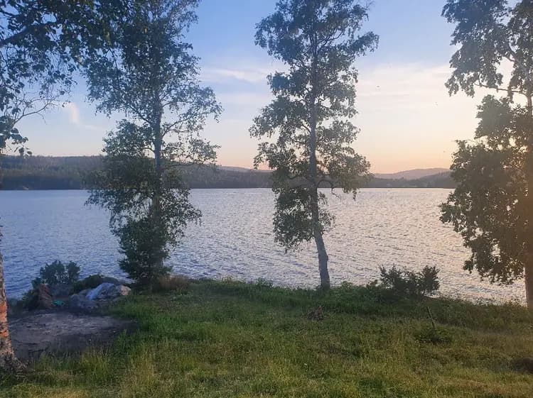 Bild av Snibbens camping som visar ett naturskönt landskap med blått vatten från en sjö i förgrunden och en horisont med skogsklädda kullar i bakgrunden.