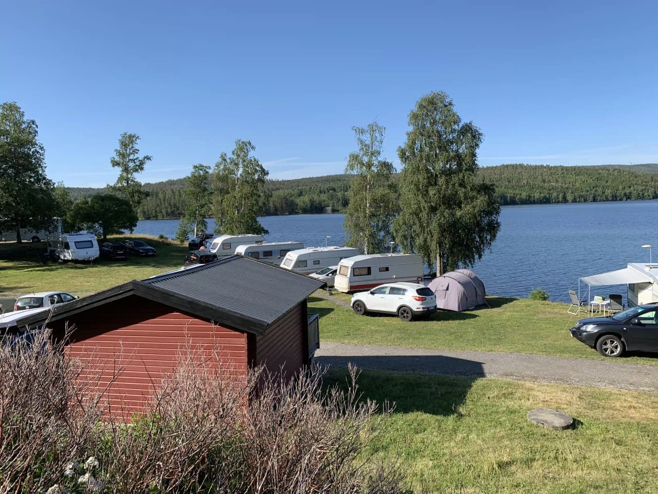 Vy över en sjö vid Snibbens Camping, omgiven av skogsklädda stränder och blå himmel i bakgrunden.