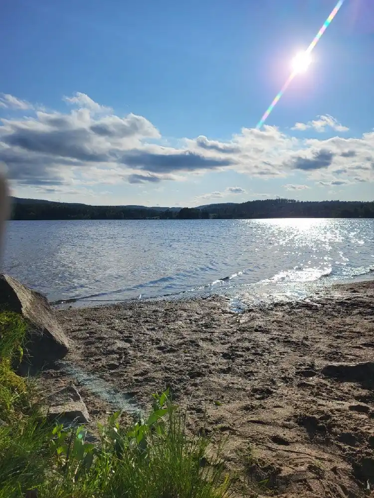 Vy över Snibbens camping med klarblå himmel och moln i bakgrunden. Framför syns ett glittrande vattenlandskap med horisonten i fjärran, vilket förstärker den natursköna känslan av platsen.