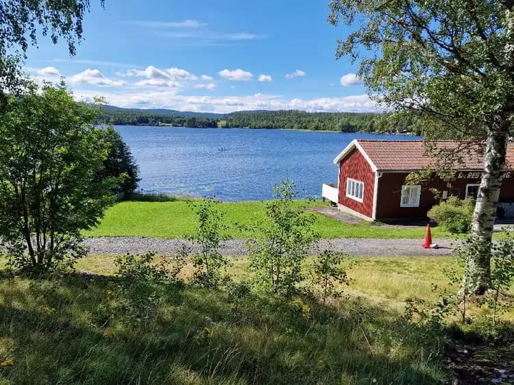 En naturskön vy över Snibbens camping med en sjö i förgrunden omgiven av grönskande växtlighet och ett litet stuga.