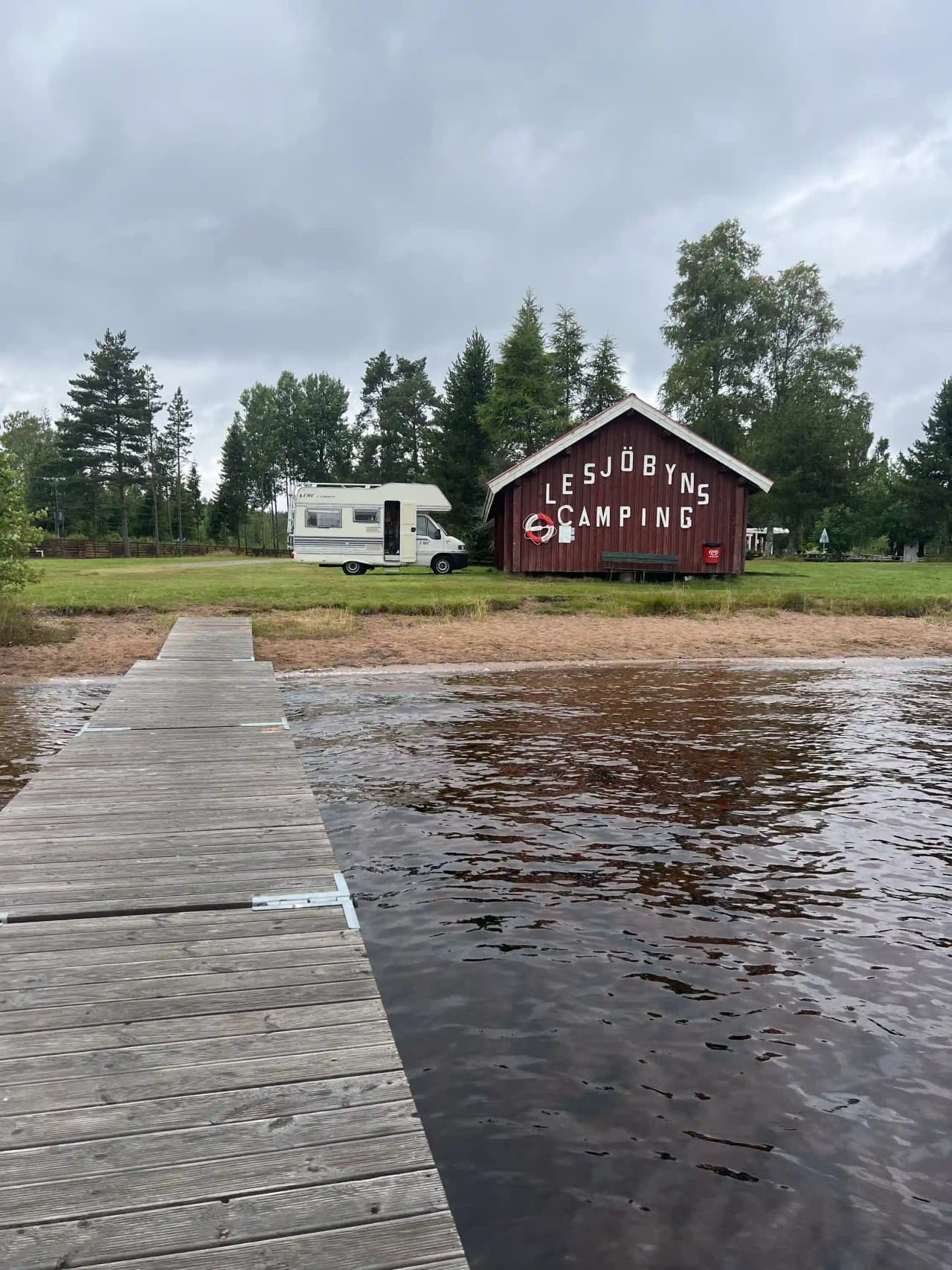 En lantlig campingplats vid Lesjöbyns camping, omgiven av en stuga och trädkantade våtmarker. En timmerstuga och ett boningshus syns i bakgrunden nära en sjöliknande vattenyta. Atmosfären är fridfull och naturskön.