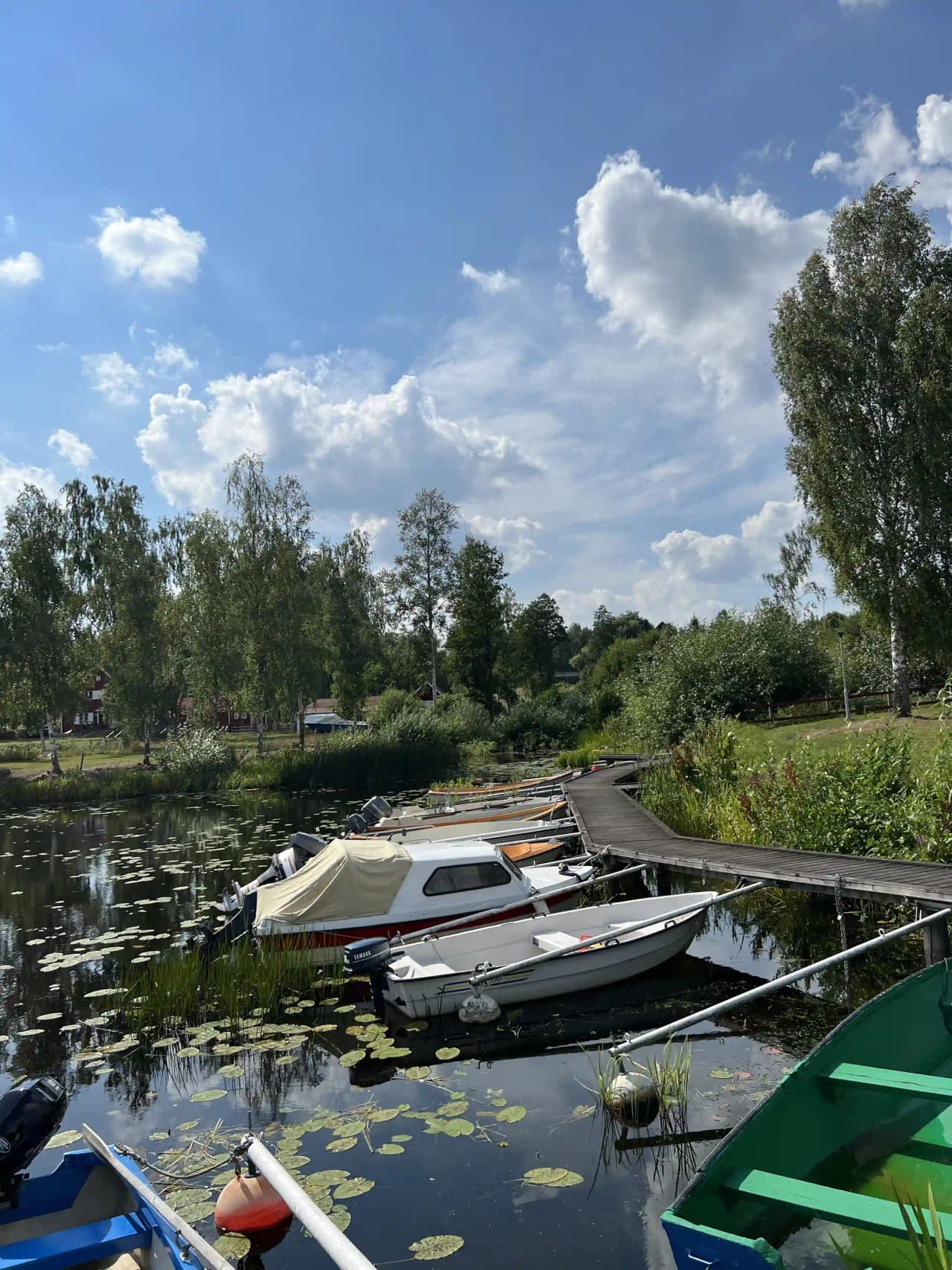 En rad båtar förtöjda vid en brygga i en lugn kanal omgiven av grönska, med speglingar i vattnet vid södra Kinda båtklubb.