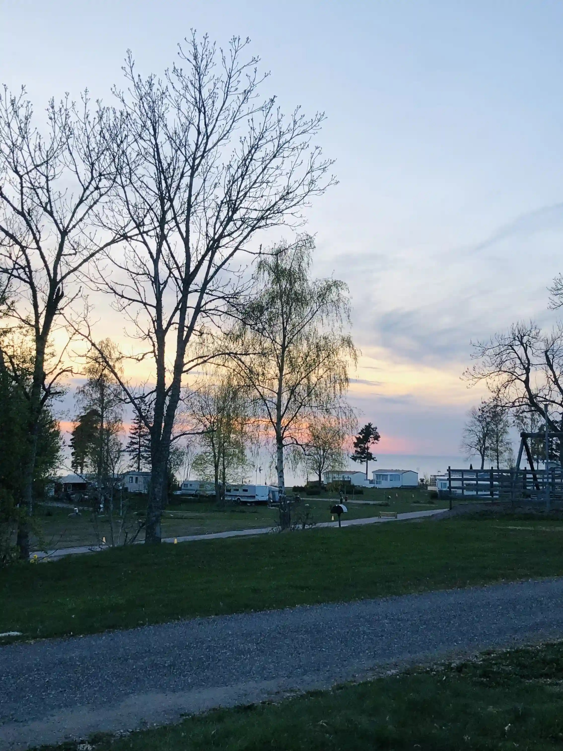 En vidsträckt vy över Askeviks camping & stugor med en klarblå himmel, omgiven av grönskande natur. Gräs och buskage bildar en naturlig ram runt det öppna landskapet medan moln seglar över horisonten i skymningen.